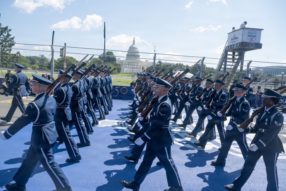 A military honor guard unit, appearing to be U.S. Air Force personnel in dress uniforms, marches in formation while carrying rifles. The ceremony takes place near the U.S. Capitol building, visible in the background, on a blue-painted surface. A tower bearing NTT IndyCar Series and 'Freedom 100' signage is visible, suggesting this is part of a racing event's opening ceremony.