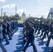 A military honor guard unit, appearing to be U.S. Air Force personnel in dress uniforms, marches in formation while carrying rifles. The ceremony takes place near the U.S. Capitol building, visible in the background, on a blue-painted surface. A tower bearing NTT IndyCar Series and 'Freedom 100' signage is visible, suggesting this is part of a racing event's opening ceremony.
