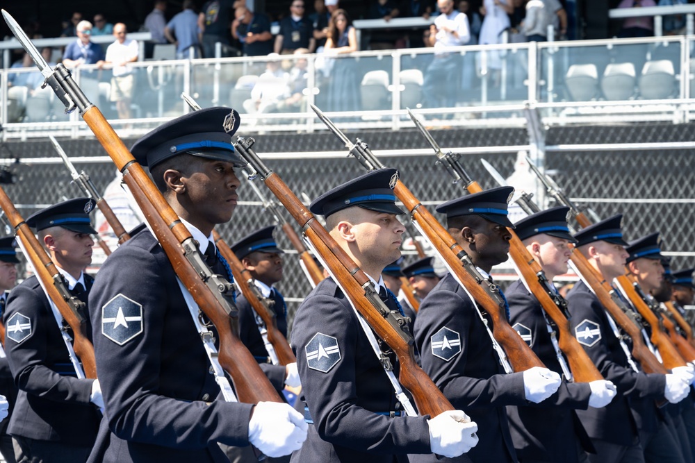 A formation of military cadets marches in a formal parade or review ceremony. They are dressed in dark navy dress uniforms with blue-banded caps and white gloves, carrying rifles with fixed bayonets at shoulder arms. Spectators are visible in bleachers in the background.