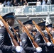 A formation of military cadets marches in a formal parade or review ceremony. They are dressed in dark navy dress uniforms with blue-banded caps and white gloves, carrying rifles with fixed bayonets at shoulder arms. Spectators are visible in bleachers in the background.