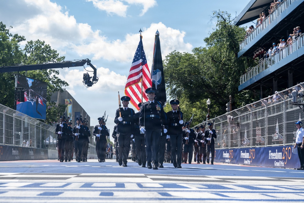 A military or law enforcement color guard is marching down a racing circuit during a pre-race ceremony for the 'Freedom 250 Grand Prix.' Personnel in dark dress uniforms carry the American flag and a POW/MIA flag along the track's start/finish straight. Spectators watch from grandstands on the right, and a camera crane is visible on the left.