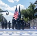 A military or law enforcement color guard is marching down a racing circuit during a pre-race ceremony for the 'Freedom 250 Grand Prix.' Personnel in dark dress uniforms carry the American flag and a POW/MIA flag along the track's start/finish straight. Spectators watch from grandstands on the right, and a camera crane is visible on the left.