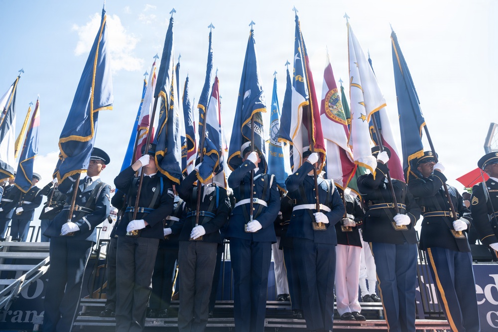 A formal military color guard ceremony featuring personnel from multiple branches of the U.S. armed forces standing at attention while holding numerous flags. The participants are dressed in distinct service dress uniforms representing different military branches, including Army, Air Force, Navy, and Marines. The ceremony appears to be a large-scale joint service event held outdoors under a partly cloudy sky.