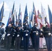 A formal military color guard ceremony featuring personnel from multiple branches of the U.S. armed forces standing at attention while holding numerous flags. The participants are dressed in distinct service dress uniforms representing different military branches, including Army, Air Force, Navy, and Marines. The ceremony appears to be a large-scale joint service event held outdoors under a partly cloudy sky.