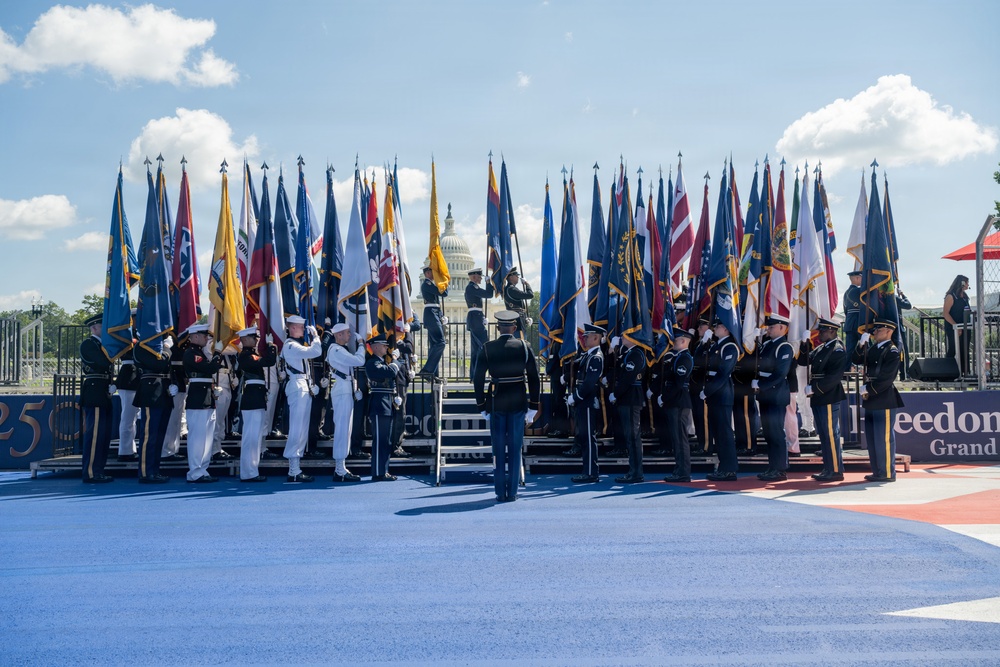 A formal military color guard ceremony is taking place on the National Mall in Washington, D.C., with the U.S. Capitol Building visible in the background. Multiple branches of the U.S. military are represented, with personnel in various dress uniforms carrying a large array of state and military flags. Partial text visible on signage suggests this may be related to a '250th' anniversary or 'Freedom' themed event.