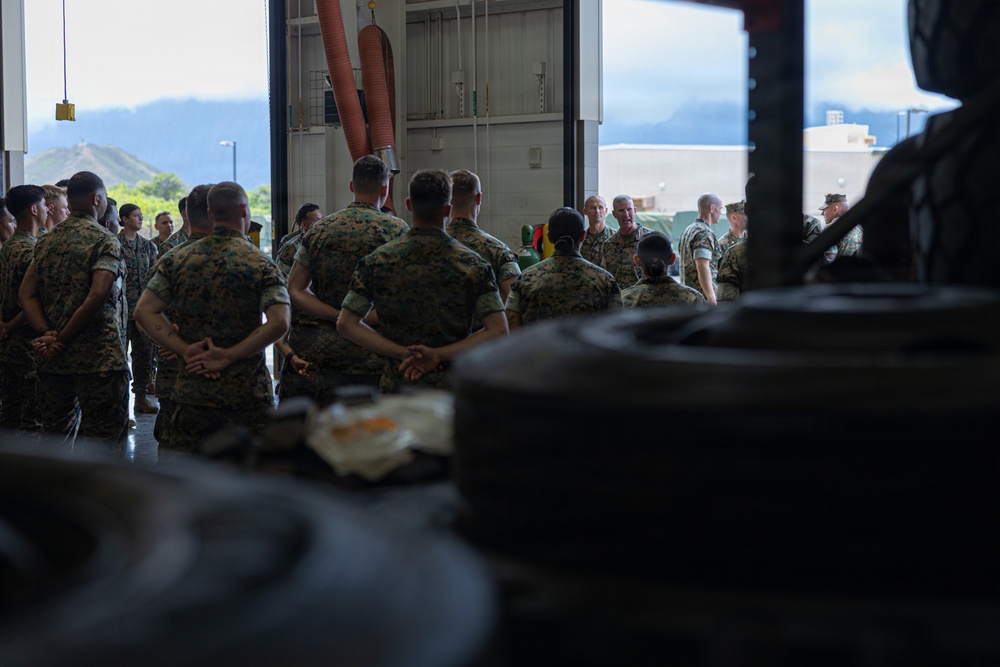 A group of military personnel in camouflage uniforms are gathered inside what appears to be a maintenance bay or hangar facility. A senior officer or official appears to be addressing the assembled troops in a briefing or formation setting. The outdoor background reveals a mountainous, tropical landscape suggesting a Pacific region installation.