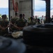 A group of military personnel in camouflage uniforms are gathered inside what appears to be a maintenance bay or hangar facility. A senior officer or official appears to be addressing the assembled troops in a briefing or formation setting. The outdoor background reveals a mountainous, tropical landscape suggesting a Pacific region installation.