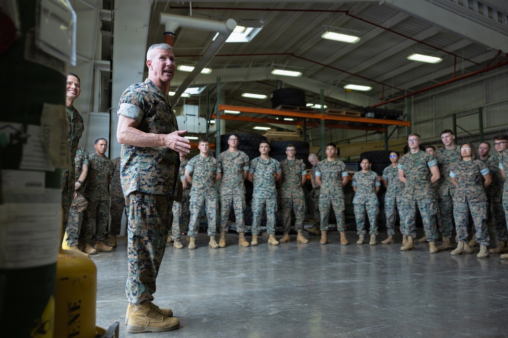 A senior military officer in U.S. Marine Corps MARPAT camouflage uniform is addressing a formation of Marines inside a large warehouse or hangar facility. The officer appears to be delivering a briefing or motivational talk to approximately 20-30 assembled Marines standing in a loose formation. The setting suggests a military installation with industrial storage infrastructure visible in the background.