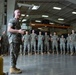 A senior military officer in U.S. Marine Corps MARPAT camouflage uniform is addressing a formation of Marines inside a large warehouse or hangar facility. The officer appears to be delivering a briefing or motivational talk to approximately 20-30 assembled Marines standing in a loose formation. The setting suggests a military installation with industrial storage infrastructure visible in the background.