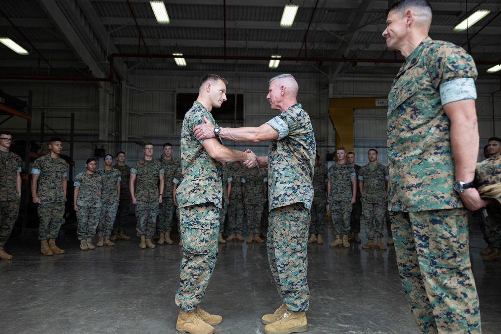 A military ceremony is taking place inside a large hangar facility where a senior officer is shaking hands with and gripping the shoulder of a junior service member, likely presenting an award or commendation. Both individuals are wearing USMC MARPAT (Marine Pattern) camouflage uniforms. A formation of Marines stands at attention in the background as witnesses to the ceremony.