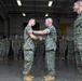 A military ceremony is taking place inside a large hangar facility where a senior officer is shaking hands with and gripping the shoulder of a junior service member, likely presenting an award or commendation. Both individuals are wearing USMC MARPAT (Marine Pattern) camouflage uniforms. A formation of Marines stands at attention in the background as witnesses to the ceremony.