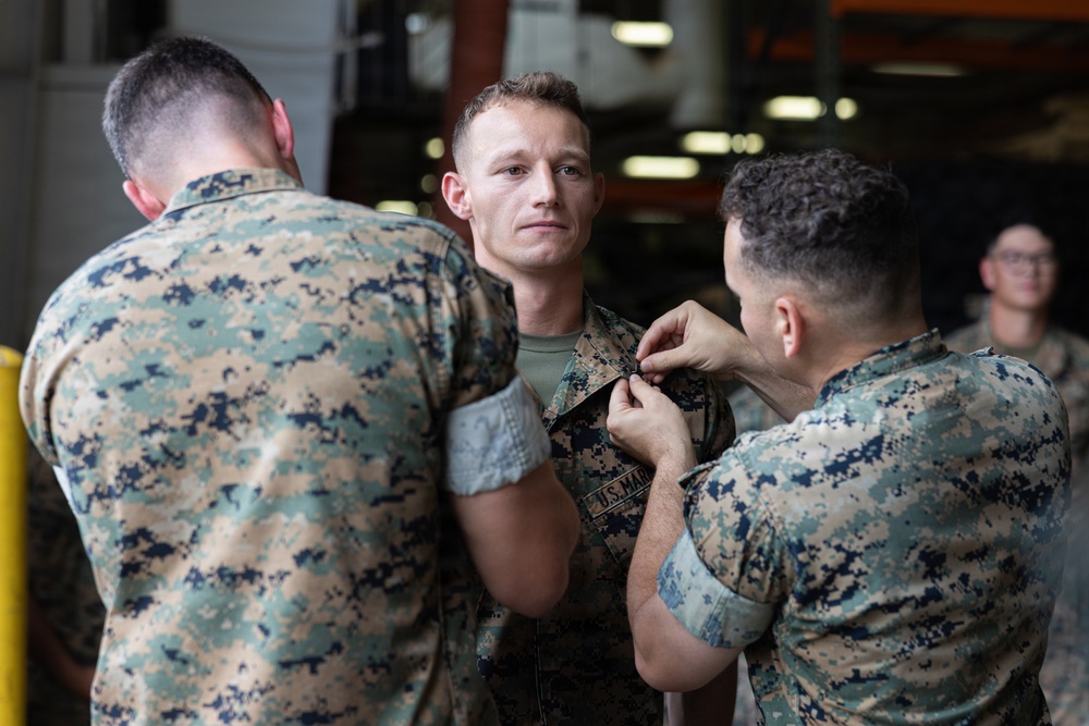 Two U.S. Marines in MARPAT digital camouflage uniforms are pinning an insignia or device onto a third Marine's collar or chest area. The central Marine faces the camera with a composed expression while the ceremony takes place. A fourth service member is visible in the background, smiling and observing the event.