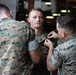 Two U.S. Marines in MARPAT digital camouflage uniforms are pinning an insignia or device onto a third Marine's collar or chest area. The central Marine faces the camera with a composed expression while the ceremony takes place. A fourth service member is visible in the background, smiling and observing the event.