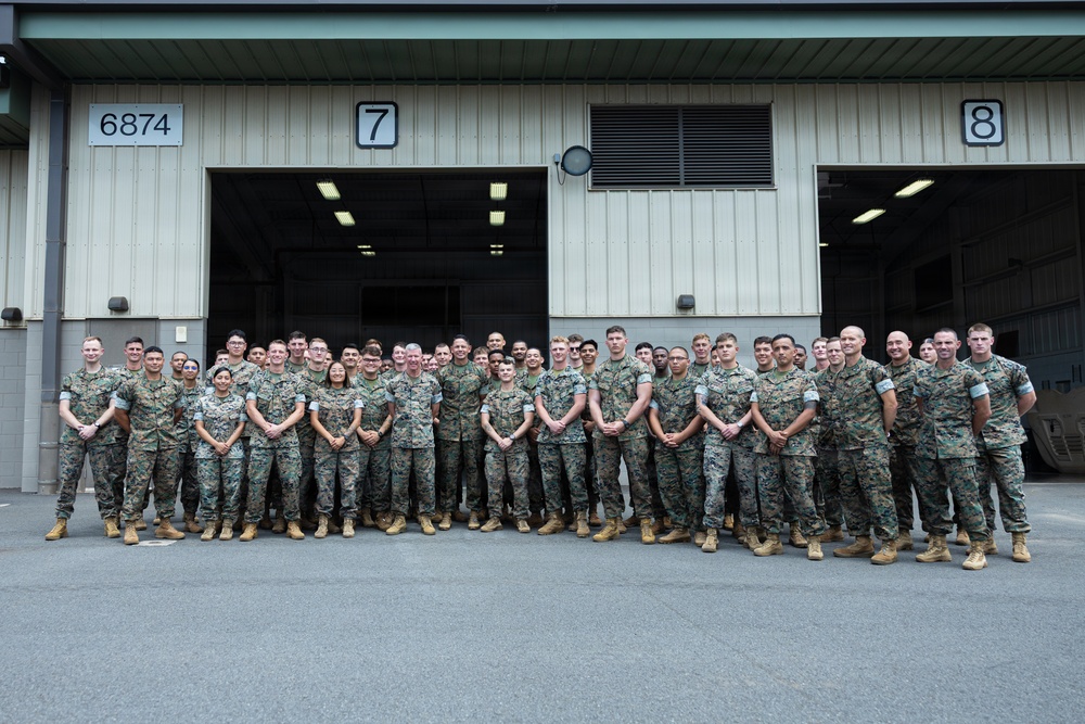 A large group of military personnel, approximately 50-60 individuals, are posed for a formal group photograph in front of a large industrial or hangar-style building numbered 6874 with bay doors labeled 7 and 8. All personnel are wearing U.S. Marine Corps MARPAT (woodland digital camouflage) uniforms with tan/coyote boots. The group includes both male and female service members arranged in two to three rows.