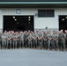 A large group of military personnel, approximately 50-60 individuals, are posed for a formal group photograph in front of a large industrial or hangar-style building numbered 6874 with bay doors labeled 7 and 8. All personnel are wearing U.S. Marine Corps MARPAT (woodland digital camouflage) uniforms with tan/coyote boots. The group includes both male and female service members arranged in two to three rows.