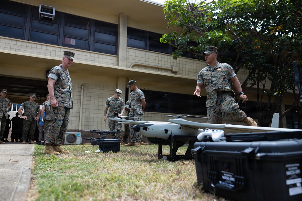 A group of U.S. Marines in woodland MARPAT camouflage uniforms are gathered outdoors near a military building, examining unmanned aerial vehicles (UAVs/drones). Two Marines in the foreground appear to be conducting a briefing or demonstration of the drone systems. Multiple UAV platforms and hard-shell transport cases are visible on the grass.