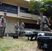 A group of U.S. Marines in woodland MARPAT camouflage uniforms are gathered outdoors near a military building, examining unmanned aerial vehicles (UAVs/drones). Two Marines in the foreground appear to be conducting a briefing or demonstration of the drone systems. Multiple UAV platforms and hard-shell transport cases are visible on the grass.