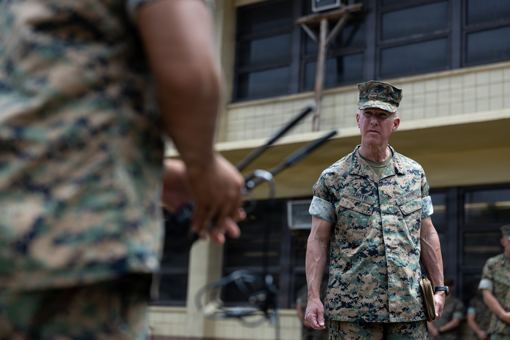 The image shows military personnel in MARPAT (Marine pattern) camouflage uniforms in what appears to be an outdoor military installation or base. A central figure with the name tape 'SMITH' is visible in focus, appearing to observe or listen during what may be a formation, briefing, or inspection. Additional personnel are visible in the background and foreground.