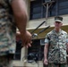 The image shows military personnel in MARPAT (Marine pattern) camouflage uniforms in what appears to be an outdoor military installation or base. A central figure with the name tape 'SMITH' is visible in focus, appearing to observe or listen during what may be a formation, briefing, or inspection. Additional personnel are visible in the background and foreground.