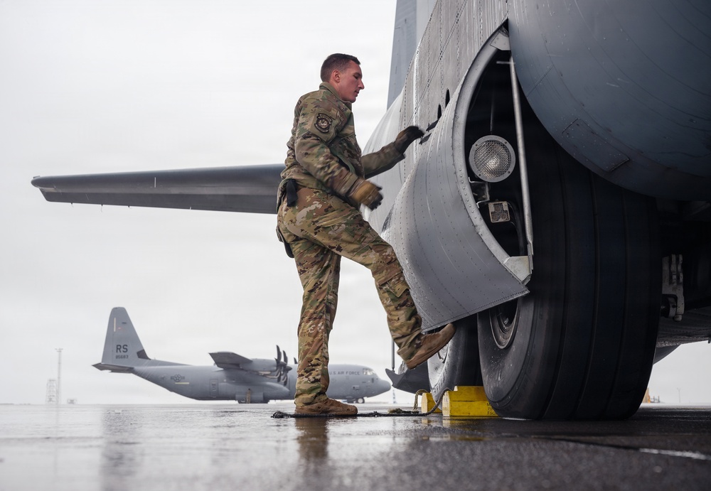 A U.S. Air Force service member in OCP (Operational Camouflage Pattern) uniform is performing a pre-flight or maintenance inspection on a large military aircraft, appearing to examine or work on the landing gear area. The wet tarmac and overcast sky suggest rainy weather conditions at a military airfield. In the background, at least two C-130 Hercules transport aircraft are visible, one bearing 'RS' tail codes indicating assignment to a specific wing.