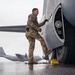 A U.S. Air Force service member in OCP (Operational Camouflage Pattern) uniform is performing a pre-flight or maintenance inspection on a large military aircraft, appearing to examine or work on the landing gear area. The wet tarmac and overcast sky suggest rainy weather conditions at a military airfield. In the background, at least two C-130 Hercules transport aircraft are visible, one bearing 'RS' tail codes indicating assignment to a specific wing.