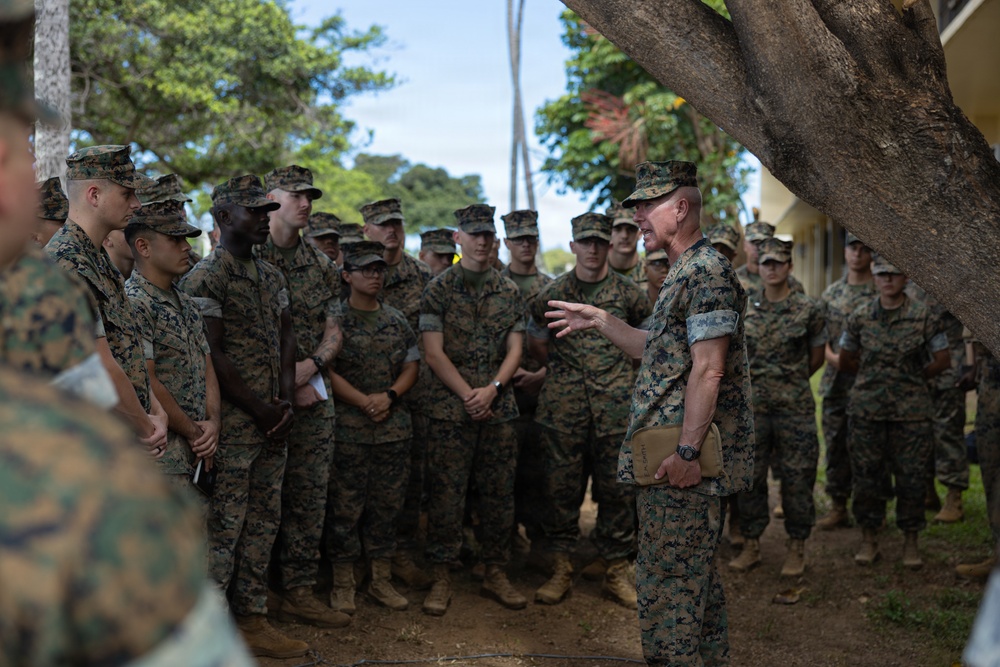 A senior military officer in U.S. Marine Corps MARPAT camouflage uniform addresses a large group of Marines gathered outdoors under trees. The speaker appears to be conducting an informal briefing or command address, gesturing with one hand while holding what appears to be a tan notebook or folder. The setting appears to be a tropical or subtropical military installation based on the vegetation visible in the background.