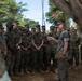 A senior military officer in U.S. Marine Corps MARPAT camouflage uniform addresses a large group of Marines gathered outdoors under trees. The speaker appears to be conducting an informal briefing or command address, gesturing with one hand while holding what appears to be a tan notebook or folder. The setting appears to be a tropical or subtropical military installation based on the vegetation visible in the background.