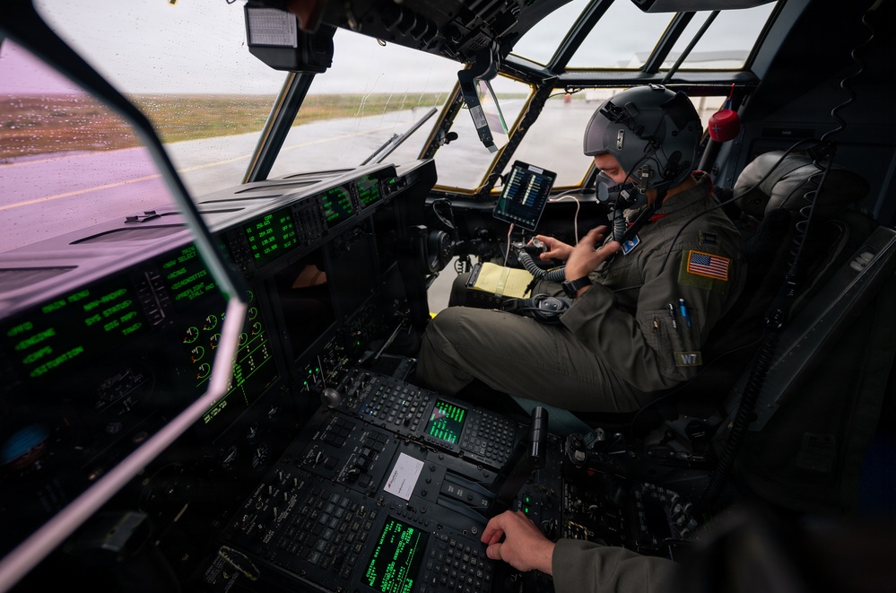 A U.S. military aviator is seated in the cockpit of a large military aircraft, likely a C-130 Hercules or similar transport aircraft, conducting pre-flight or ground operations. The pilot is wearing a flight suit, helmet, and oxygen mask while interacting with a handheld device and cockpit controls. The aircraft appears to be stationary on a wet tarmac, visible through rain-streaked windows.