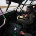 A U.S. military aviator is seated in the cockpit of a large military aircraft, likely a C-130 Hercules or similar transport aircraft, conducting pre-flight or ground operations. The pilot is wearing a flight suit, helmet, and oxygen mask while interacting with a handheld device and cockpit controls. The aircraft appears to be stationary on a wet tarmac, visible through rain-streaked windows.