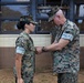 Two U.S. Marine Corps personnel in MARPAT woodland camouflage uniforms are engaged in what appears to be a military decoration or award ceremony outdoors. A senior male Marine is pinning something onto the uniform of a female Marine, who stands at attention. A third uniformed individual is partially visible in the background.