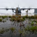 A military turboprop aircraft, consistent with a C-130 Hercules variant, sits on a wet flight line apron with spinning propellers indicating active engines. Two personnel in camouflage uniforms are visible near the aircraft conducting ground operations. The low-angle shot captures standing water and tufts of grass in the foreground, with a large aircraft hangar visible in the background under overcast skies.