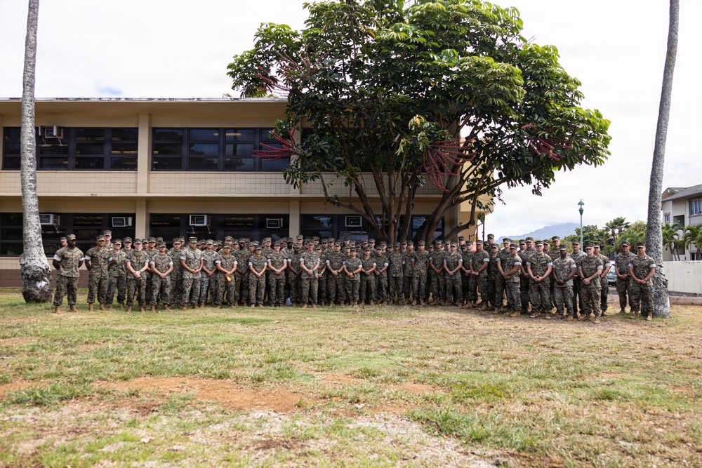 A large group of military personnel in camouflage uniforms are posed for a formal group photograph outdoors on a military installation. They are standing in a single extended row on a grassy area in front of a tan multi-story building. The tropical vegetation, including palm trees and a large broadleaf tree, suggests a location in Hawaii or a similar tropical region.