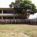 A large group of military personnel in camouflage uniforms are posed for a formal group photograph outdoors on a military installation. They are standing in a single extended row on a grassy area in front of a tan multi-story building. The tropical vegetation, including palm trees and a large broadleaf tree, suggests a location in Hawaii or a similar tropical region.