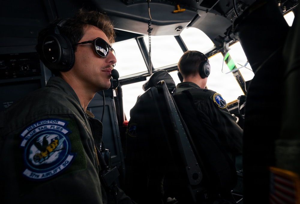 Two military aircrew members are seated in the cockpit of a large military transport aircraft, likely a C-130 Hercules, during flight. The foreground pilot wears Bose headphones, reflective aviator sunglasses, and a flight suit with a patch reading 'INSTRUCTOR - BLUE TAIL FLIES - 37TH AIRLIFT SQ.' The image captures an in-flight operational scene with overcast skies visible through the cockpit windows.