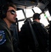 Two military aircrew members are seated in the cockpit of a large military transport aircraft, likely a C-130 Hercules, during flight. The foreground pilot wears Bose headphones, reflective aviator sunglasses, and a flight suit with a patch reading 'INSTRUCTOR - BLUE TAIL FLIES - 37TH AIRLIFT SQ.' The image captures an in-flight operational scene with overcast skies visible through the cockpit windows.