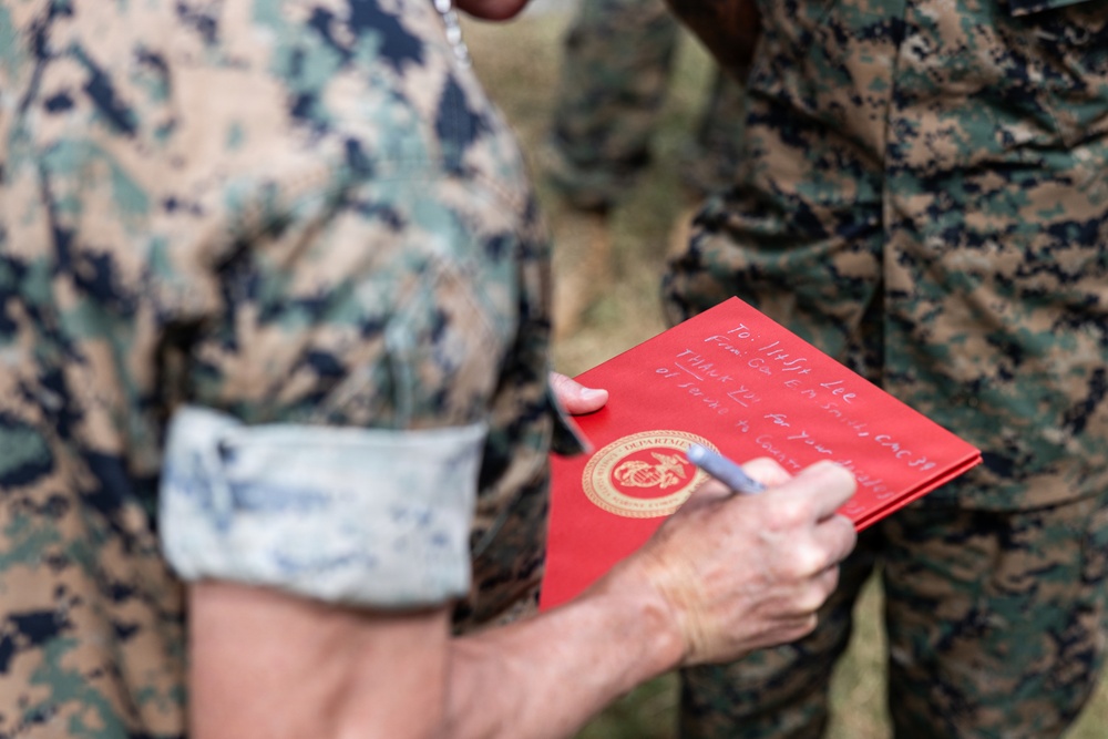A person in U.S. Marine Corps MARPAT camouflage uniform is writing a personal inscription on a red official Department of the Navy/Marine Corps folder or certificate cover. The red folder bears a gold official seal consistent with the Department of the Navy or Marine Corps emblem. The handwritten inscription appears to read 'To [name] Lee, From Gen [name], Thank You for your [service/dedication] of [years] to [Corps/country].'