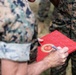 A person in U.S. Marine Corps MARPAT camouflage uniform is writing a personal inscription on a red official Department of the Navy/Marine Corps folder or certificate cover. The red folder bears a gold official seal consistent with the Department of the Navy or Marine Corps emblem. The handwritten inscription appears to read 'To [name] Lee, From Gen [name], Thank You for your [service/dedication] of [years] to [Corps/country].'