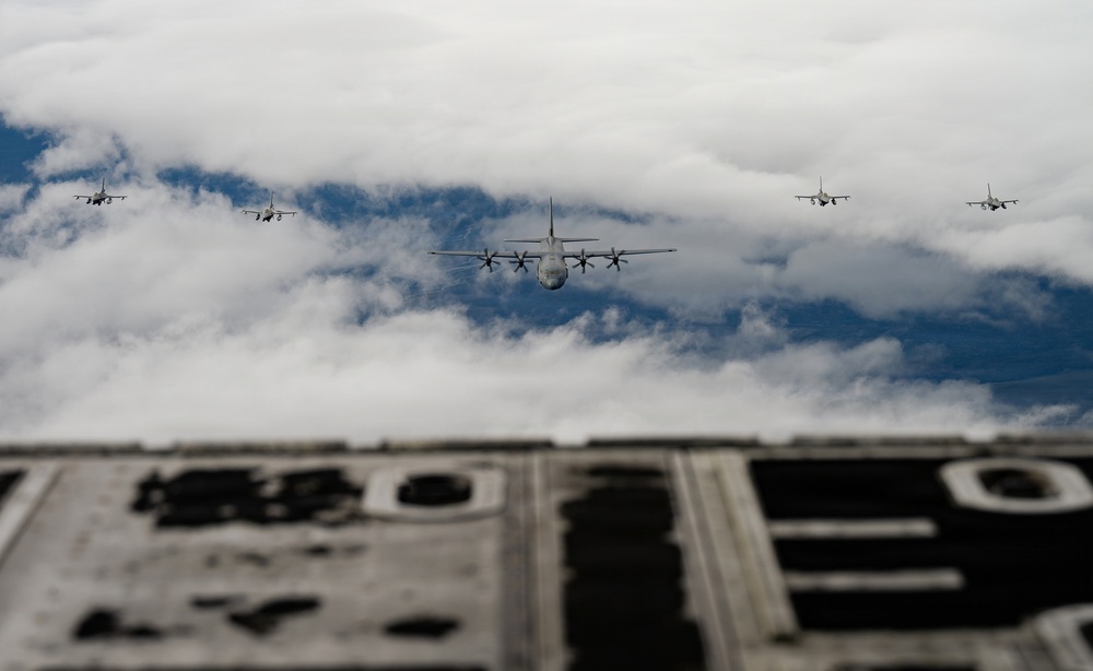 A military formation flight photographed from the rear ramp or fuselage of a large transport aircraft. The formation consists of one large four-engine turboprop transport aircraft in the center, flanked by four smaller jet fighters in a symmetrical escort formation. The image is taken from an elevated or inverted perspective, with partial view of the photographing aircraft's structure in the foreground.