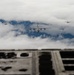 A military formation flight photographed from the rear ramp or fuselage of a large transport aircraft. The formation consists of one large four-engine turboprop transport aircraft in the center, flanked by four smaller jet fighters in a symmetrical escort formation. The image is taken from an elevated or inverted perspective, with partial view of the photographing aircraft's structure in the foreground.