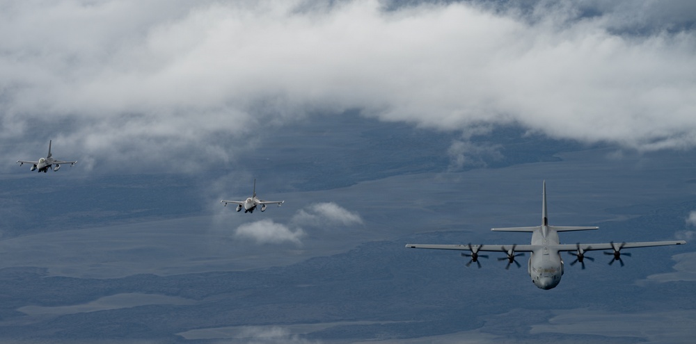 Three military aircraft are flying in formation over a vast, partially cloud-covered landscape. The formation consists of one large turboprop transport aircraft in the foreground and two smaller jet fighters trailing behind. The scene appears to be a military exercise or escort mission photographed from another airborne platform.