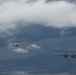Three military aircraft are flying in formation over a vast, partially cloud-covered landscape. The formation consists of one large turboprop transport aircraft in the foreground and two smaller jet fighters trailing behind. The scene appears to be a military exercise or escort mission photographed from another airborne platform.