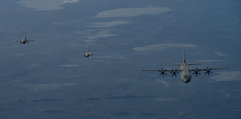 Three military aircraft are flying in formation over a vast, arid landscape viewed from altitude. The formation consists of one large four-engine turboprop transport aircraft on the right and two smaller single-engine jet fighters positioned to the left at varying distances. The terrain below appears to be a semi-arid or desert region with scattered water bodies or dry lake beds visible.