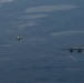 Three military aircraft are flying in formation over a vast, arid landscape viewed from altitude. The formation consists of one large four-engine turboprop transport aircraft on the right and two smaller single-engine jet fighters positioned to the left at varying distances. The terrain below appears to be a semi-arid or desert region with scattered water bodies or dry lake beds visible.