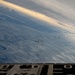 An aerial formation flight photographed from the open rear ramp of a large military transport aircraft, showing multiple aircraft flying in loose formation over a snow-covered, glaciated landscape. The image captures at least five aircraft including what appear to be C-130 Hercules turboprop transports and smaller jet fighters flying in echelon. The terrain below shows extensive snow and ice coverage with mountain ranges visible in the background under partly cloudy skies.