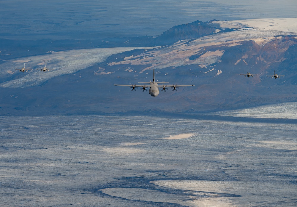 A military formation flight consisting of one large turboprop transport aircraft flanked by four fighter jets flying over a snow-covered and glaciated arctic or subarctic landscape. The terrain below features extensive glaciers, ice fields, and rugged mountain ridges with partial snow cover. The image appears to be taken from another aircraft flying ahead of or above the formation.