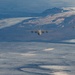 A military formation flight consisting of one large turboprop transport aircraft flanked by four fighter jets flying over a snow-covered and glaciated arctic or subarctic landscape. The terrain below features extensive glaciers, ice fields, and rugged mountain ridges with partial snow cover. The image appears to be taken from another aircraft flying ahead of or above the formation.