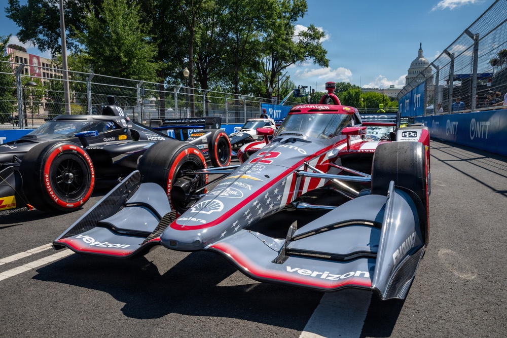 The image shows multiple IndyCar racing cars lined up on a street circuit, with the United States Capitol building visible in the background. The foreground features a prominently displayed car with Verizon sponsorship and an American flag-themed livery in gray, red, and white. This appears to be the NTT IndyCar Series race held in Washington D.C.