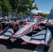 The image shows multiple IndyCar racing cars lined up on a street circuit, with the United States Capitol building visible in the background. The foreground features a prominently displayed car with Verizon sponsorship and an American flag-themed livery in gray, red, and white. This appears to be the NTT IndyCar Series race held in Washington D.C.