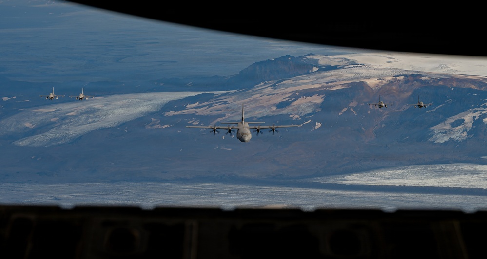 The image is taken from inside a military aircraft looking out through what appears to be a rear cargo door or ramp opening. A formation of military aircraft, including a large four-engine turboprop transport (likely a C-130 Hercules) in the center and multiple fighter jets (likely F-16s) flanking on both sides, are flying over a snow-capped mountainous terrain. The landscape below features glaciers, rocky ridgelines, and appears consistent with Arctic or sub-Arctic geography such as Iceland or Greenland.