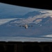 The image is taken from inside a military aircraft looking out through what appears to be a rear cargo door or ramp opening. A formation of military aircraft, including a large four-engine turboprop transport (likely a C-130 Hercules) in the center and multiple fighter jets (likely F-16s) flanking on both sides, are flying over a snow-capped mountainous terrain. The landscape below features glaciers, rocky ridgelines, and appears consistent with Arctic or sub-Arctic geography such as Iceland or Greenland.